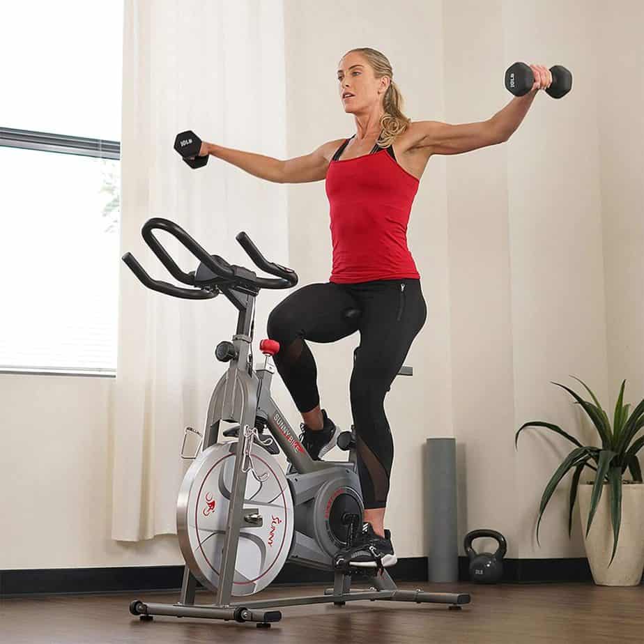 A lady workouts with the Sunny Health & Fitness Synergy SF-B1879 Indoor Cycling Bike while using dumbbells 